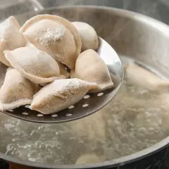 Gotowanie pierogów: na łyżce cedzakowej świeżo uformowane pierogi czekają na wrzucenie do wrzątku. W garnku bulgocze woda, gotowe do przyjęcia pysznego farszu.