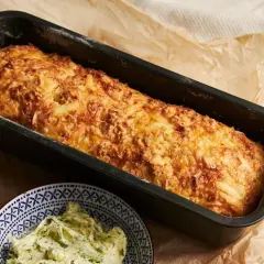 Chrupiąca babka ziemniaczana w keksówce, obok miseczka z masłem ziołowym.