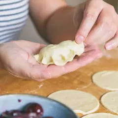 Dłonie formują pieroga z ciasta na pierogi bez jajka. Na blacie leżą okrągłe placki z ciasta.