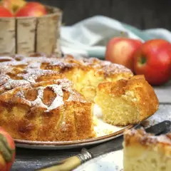 Wilgotne ciasto z jabłkami na oleju, posypane cukrem pudrem, kusi apetycznym wyglądem. Obok leżą świeże jabłka.