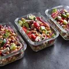 Trzy szklane pojemniki wypełnione kolorową sałatką z ciecierzycą, ogórkiem, pomidorem, czerwoną cebulą i fetą.