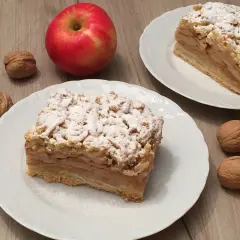 Szybka szarlotka z jabłkami ze słoika, posypana cukrem pudrem, obok orzechy i jabłko.