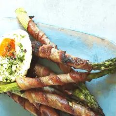 Pieczone szparagi w boczku z jajkiem sadzonym na niebieskiej desce.
