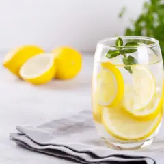Szklanka wody z plasterkami cytryny i miętą, na tle cytryn i zielonych liści.