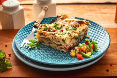 Kawałek pysznej, warzywnej lasagne na niebieskim talerzu, posypany natką i pieprzem. To bardzo dobry przepis na lazanię, który zachwyci każdego!