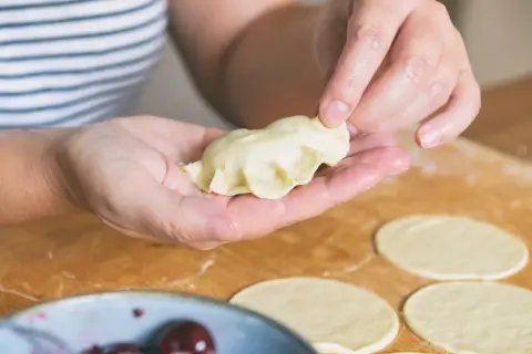 Dłonie formują pieroga z ciasta na pierogi bez jajka. Na blacie leżą okrągłe placki z ciasta.