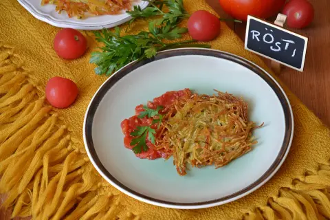 Chrupiące rosti ziemniaczane z pomidorami i natką pietruszki na talerzu, obok tabliczka z napisem "Rösti".