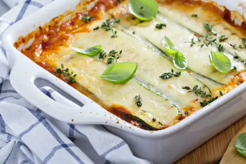 Pyszne, zapiekane w białym naczyniu danie, gdzie cienkie plastry cukinii zastępują makaron w lasagne. Całość polana sosem pomidorowym i posypana serem, udekorowana listkami bazylii i tymianku.