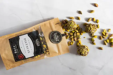 Opakowanie pistacji i trzy praliny z pistacjami rozsypane na marmurowym blacie. Warto sprawdzić pistacje kcal przed spożyciem.
