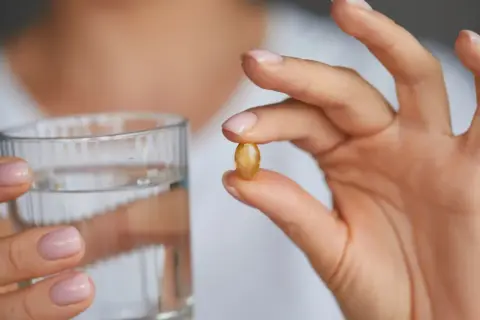 Dłoń trzyma kapsułkę omega 3 i szklankę wody. Kiedy brać omega 3? Najlepiej z posiłkiem.