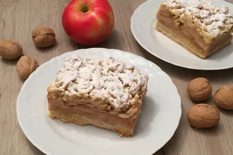 Szybka szarlotka z jabłkami ze słoika, posypana cukrem pudrem, obok orzechy i jabłko.