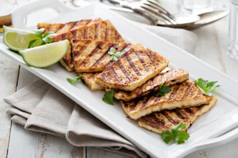 Grillowany ser halloumi, idealny jako marynata na grilla, podany z ćwiartkami limonki i natką pietruszki na białej tacy.