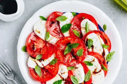 Sałatka caprese: plastry soczystego pomidora z mozzarellą, skropione balsamico i udekorowane listkami bazylii.