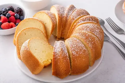 Pyszna babka piaskowa, jak z przepisu babci, posypana cukrem pudrem, z owocami i widelcami obok.