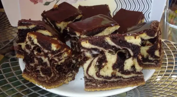 Wilgotne ciasto zebra z polewą czekoladową, pokrojone na kwadratowe kawałki, prezentuje się apetycznie na białym talerzu.