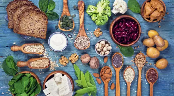 Różnorodne produkty roślinne: pieczywo, warzywa, nasiona, orzechy, fasola, tofu, na niebieskim tle.