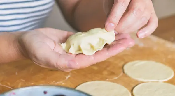 Dłonie formują pieroga z ciasta na pierogi bez jajka. Na blacie leżą okrągłe placki z ciasta.