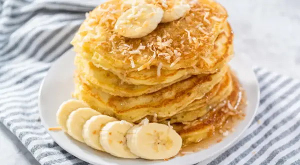 Stos puszystych naleśników z mąki kokosowej, udekorowany plasterkami banana i wiórkami kokosowymi, polany syropem.