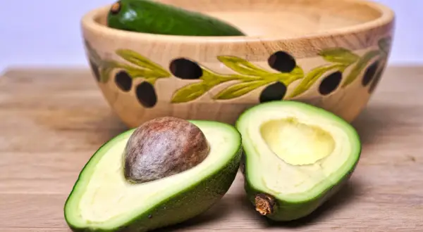 Dwa połówki awokado, jedno z pestką, leżą na drewnianym blacie obok ozdobnej misy. Zastanawiasz się nad awokado kcal?