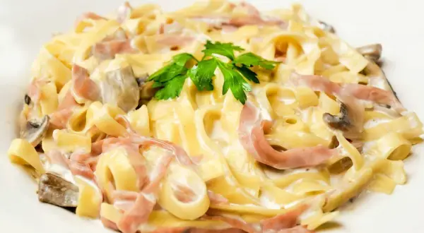 Tagliatelle z szynką i pieczarkami w kremowym sosie, z dodatkiem mozzarelli. Udekorowane natką pietruszki.