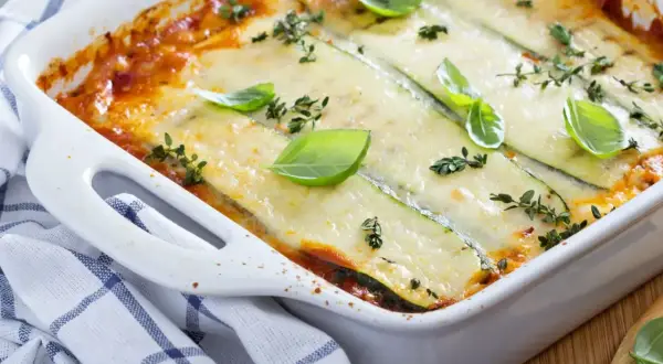 Pyszne, zapiekane w białym naczyniu danie, gdzie cienkie plastry cukinii zastępują makaron w lasagne. Całość polana sosem pomidorowym i posypana serem, udekorowana listkami bazylii i tymianku.