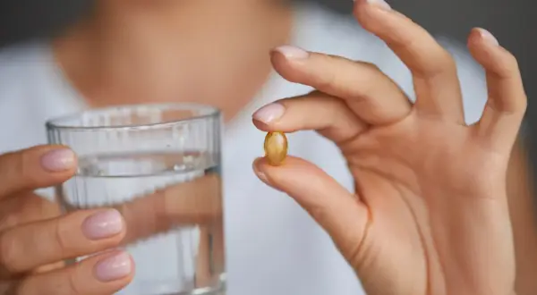 Dłoń trzyma kapsułkę omega 3 i szklankę wody. Kiedy brać omega 3? Najlepiej z posiłkiem.