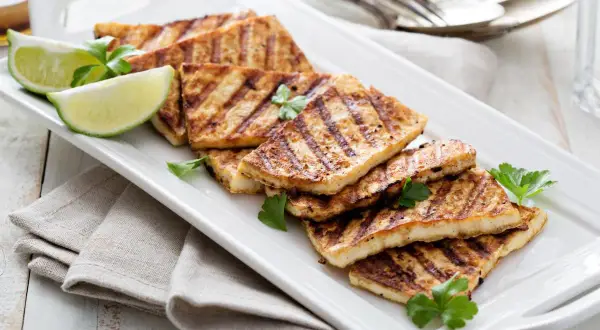 Grillowany ser halloumi, idealny jako marynata na grilla, podany z ćwiartkami limonki i natką pietruszki na białej tacy.