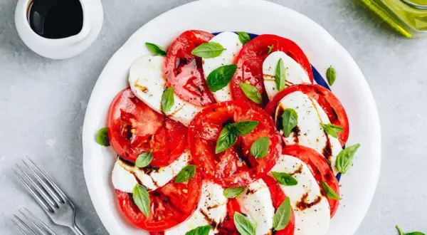 Sałatka caprese: plastry soczystego pomidora z mozzarellą, skropione balsamico i udekorowane listkami bazylii.