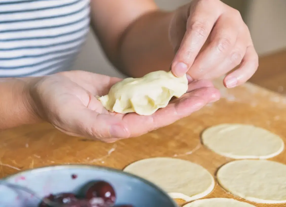 Dłonie formują pieroga z ciasta na pierogi bez jajka. Na blacie leżą okrągłe placki z ciasta.