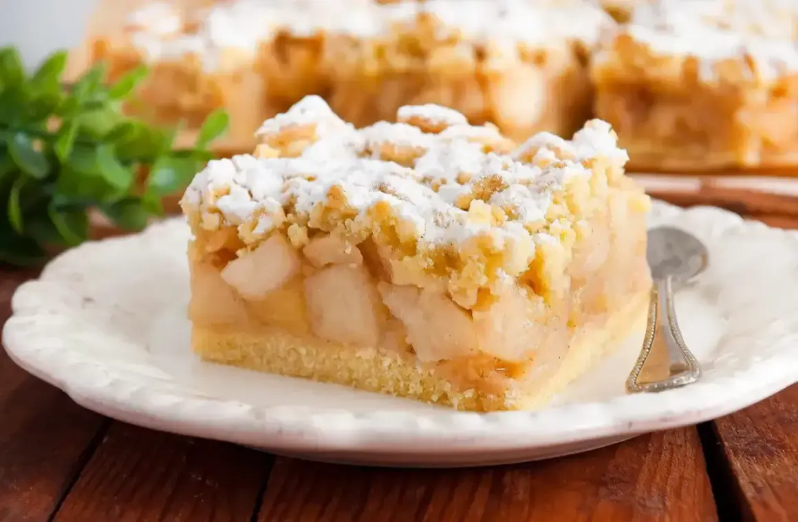 Kawałek pysznego ciasta jabłkowego z kruszonką, posypany cukrem pudrem. Idealny przykład, jak zrobić kruche ciasto.