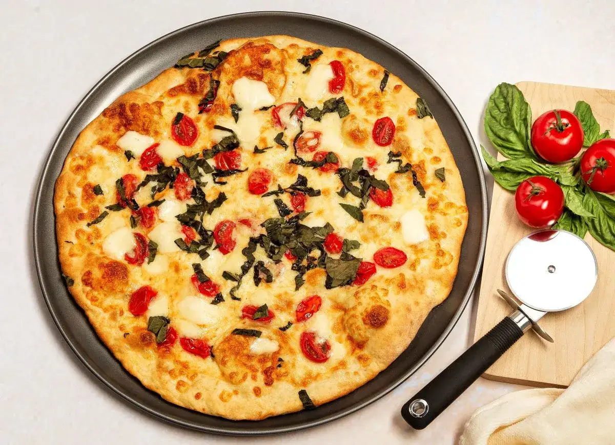 Pyszna pizza z serka wiejskiego, pomidorkami koktajlowymi i bazylią. Obok leży krajalnica do pizzy i świeże pomidory.