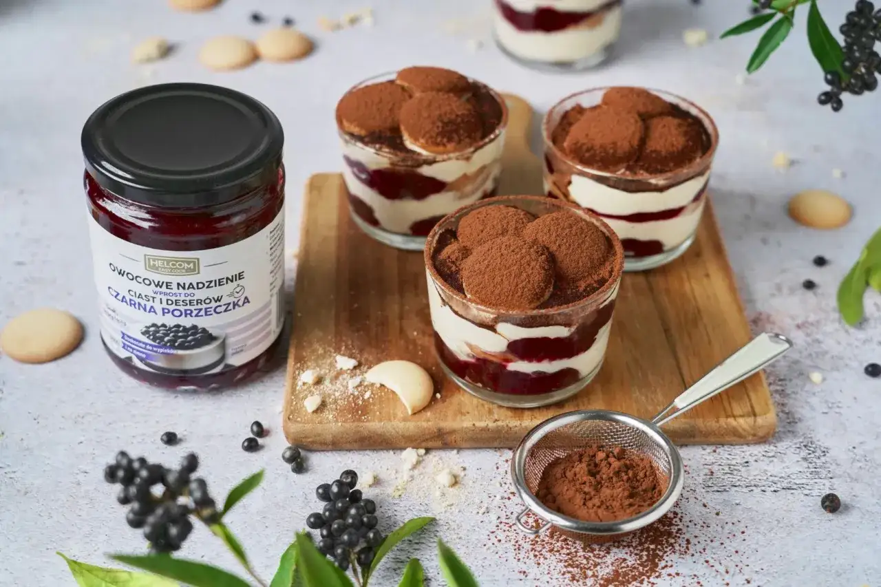 Najprostsze tiramisu z czarną porzeczką w słoiczkach, ozdobione biszkoptami i kakao. Obok słoik dżemu.