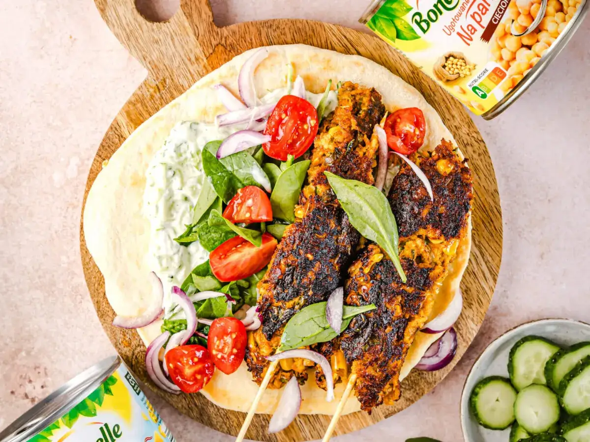 Pyszny kebab domowy na desce z tortillą, mięsem, warzywami i sosem. Obok puszka ciecierzycy i plasterki ogórka.