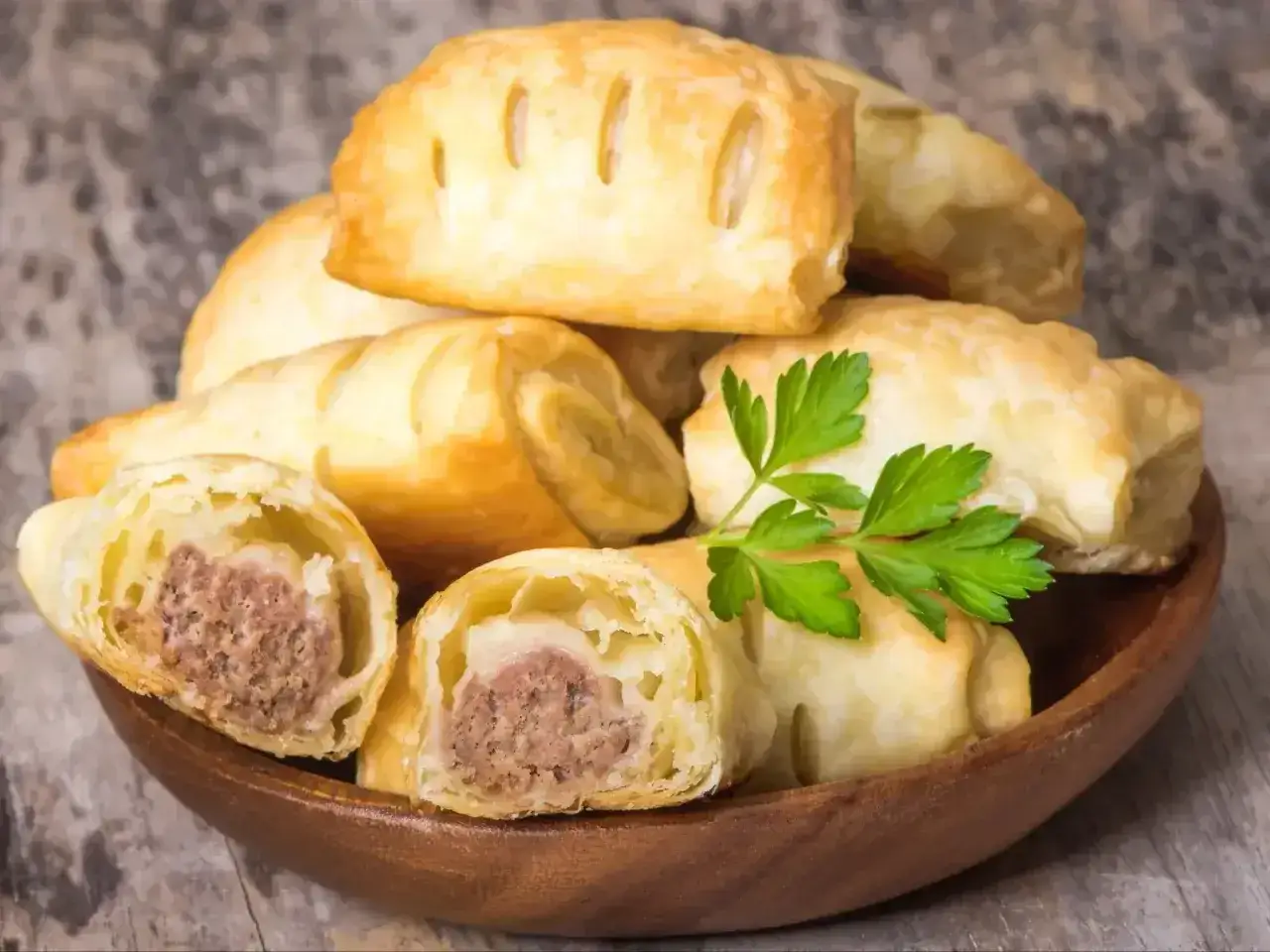 Stos pieczonego ciasta francuskiego z kiełbaską w środku, ozdobiony gałązką pietruszki.