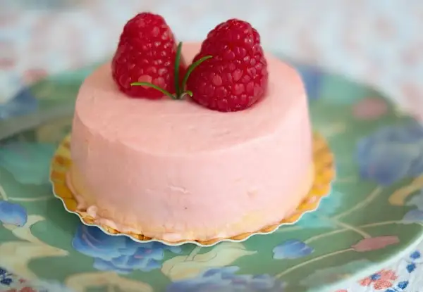Delikatny krem malinowy z mascarpone do tortu, ozdobiony dwoma świeżymi malinami i gałązką rozmarynu, na zielonym talerzyku w kwiaty.