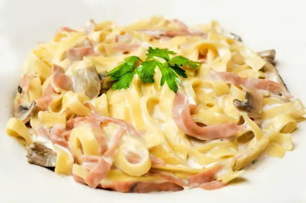Tagliatelle z szynką i pieczarkami w kremowym sosie, z dodatkiem mozzarelli. Udekorowane natką pietruszki.