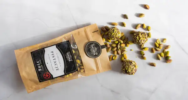 Opakowanie pistacji i trzy praliny z pistacjami rozsypane na marmurowym blacie. Warto sprawdzić pistacje kcal przed spożyciem.
