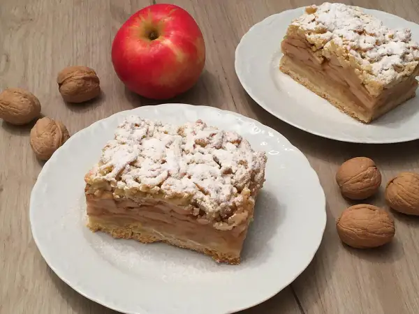 Szybka szarlotka z jabłkami ze słoika, posypana cukrem pudrem, obok orzechy i jabłko.