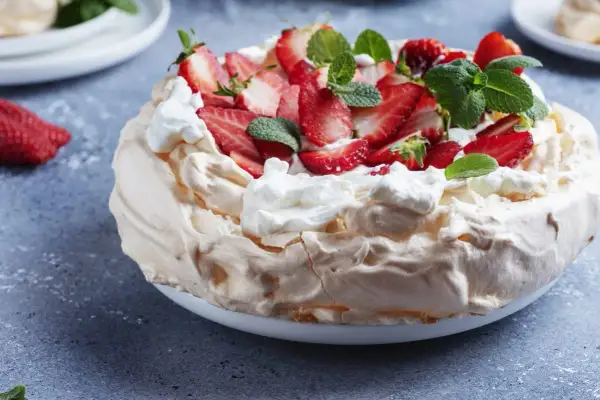Puszysty tort bezowy z mascarpone i bitą śmietaną, ozdobiony świeżymi truskawkami i miętą.