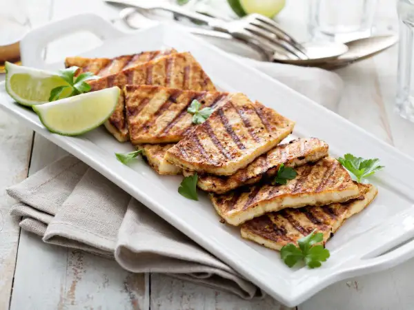 Grillowany ser halloumi, idealny jako marynata na grilla, podany z ćwiartkami limonki i natką pietruszki na białej tacy.